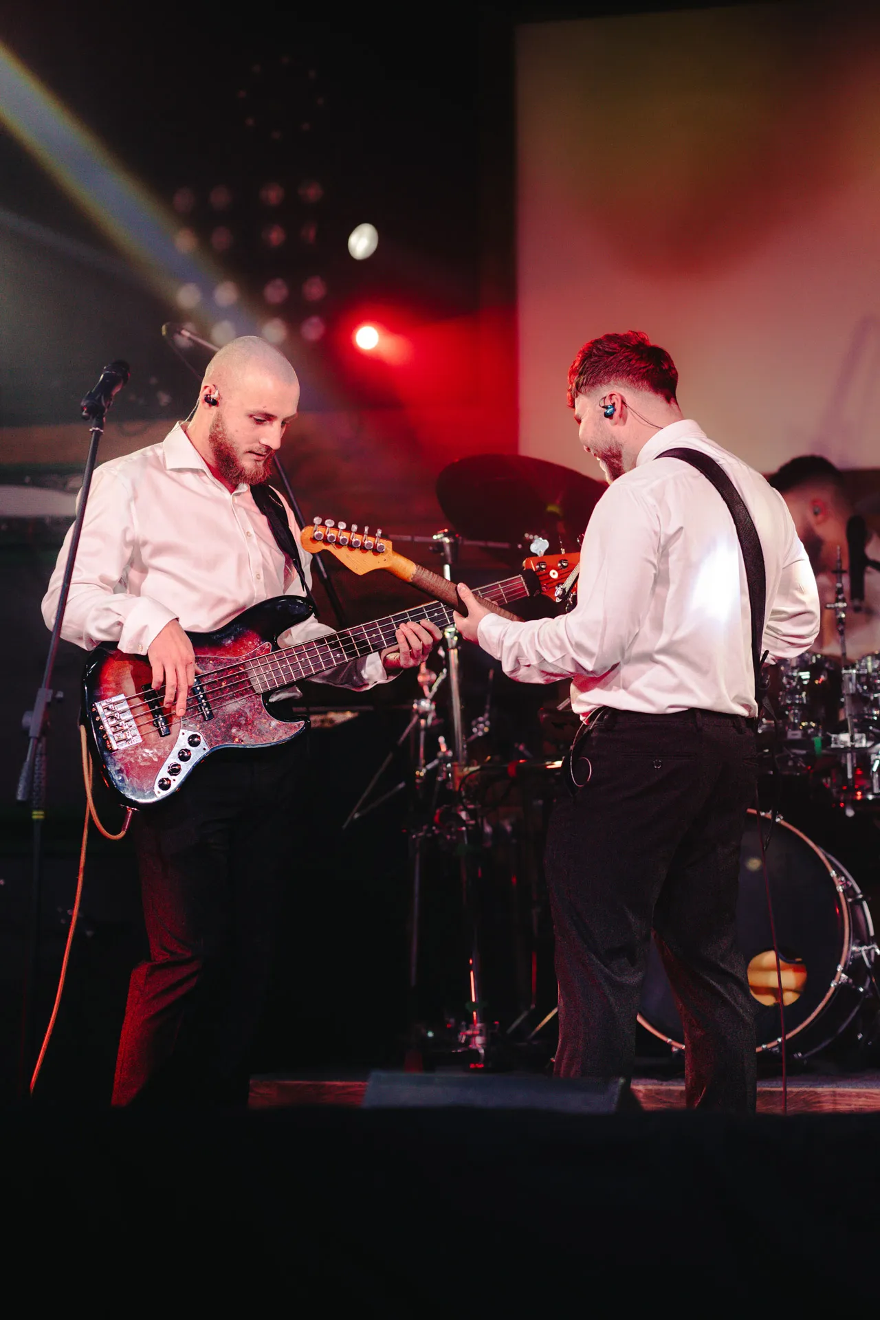 Two musicians wearing white shirts playing electric guitars on stage with red and white lighting.