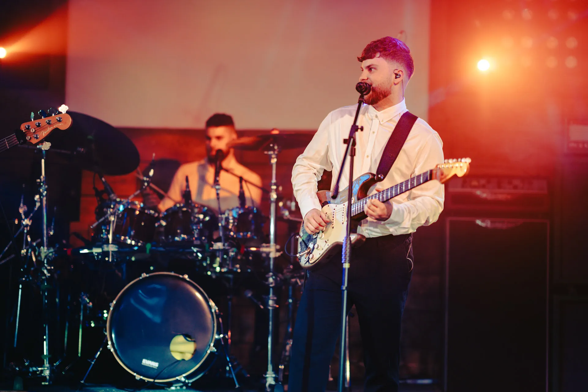Male guitarist singing into a microphone with a drummer playing in the background on stage.