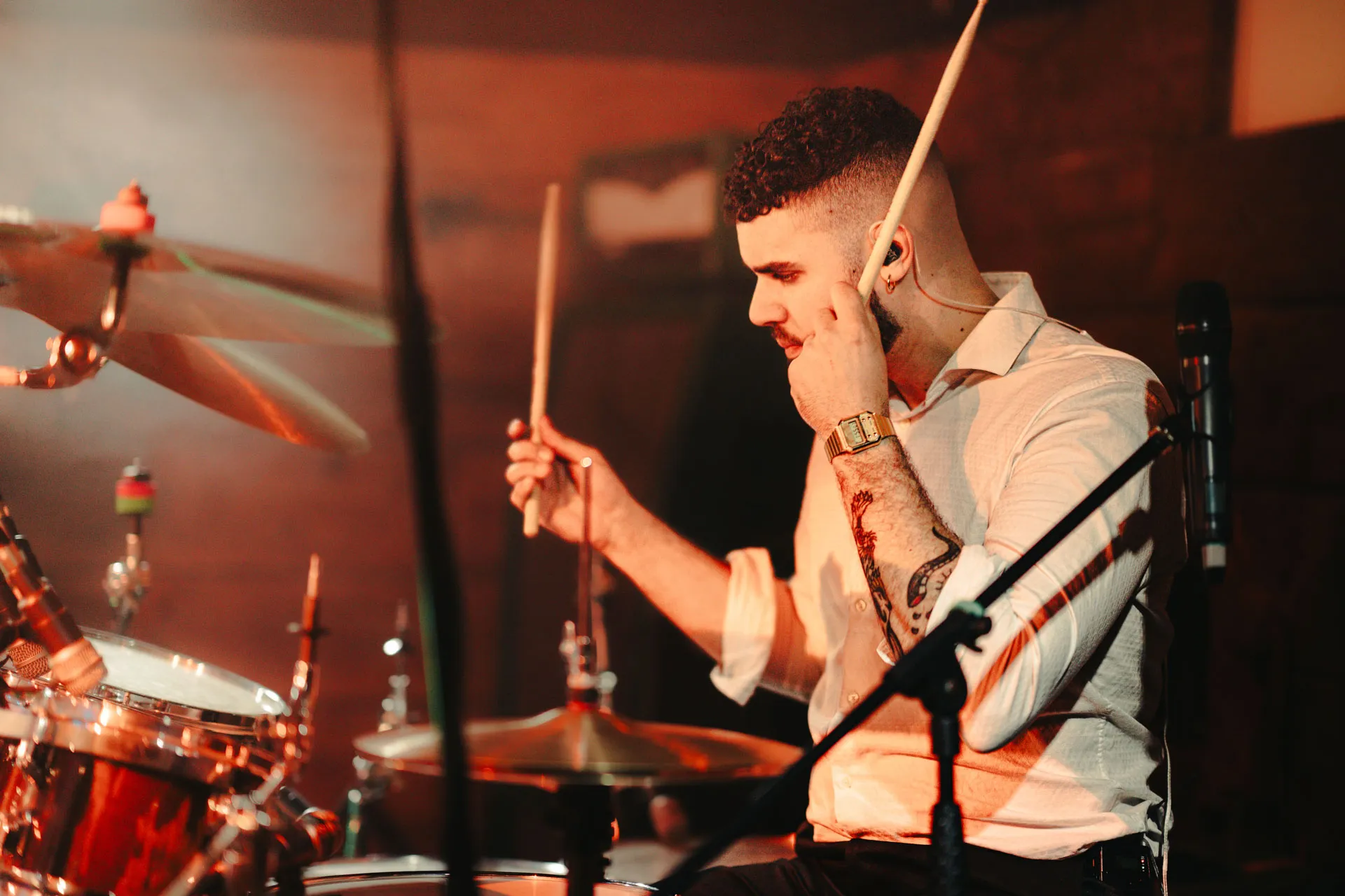 Man with tattoo playing drums holding drumsticks in a dimly lit setting.