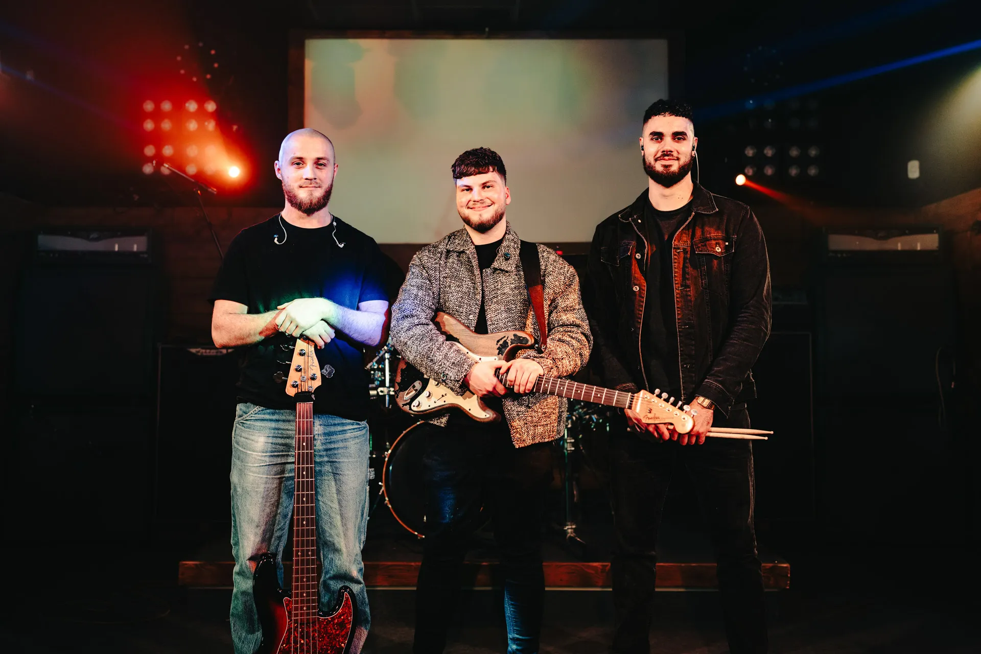 Three male band members posing with instruments on a dimly lit stage with colorful lights.