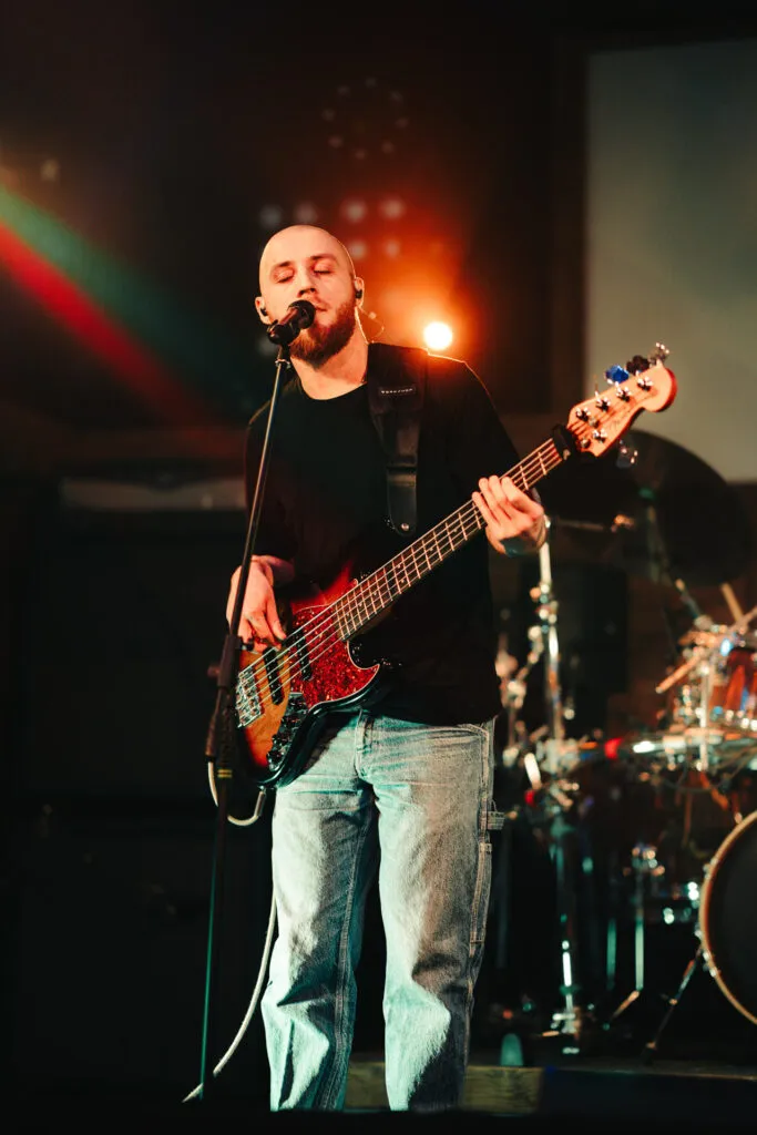 Man with a beard playing electric bass guitar and singing into a microphone on stage.